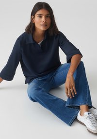 Haut style polo bleu marine en tissu lisse avec un col, associé à un jean évasé de lavage moyen et des baskets blanches.