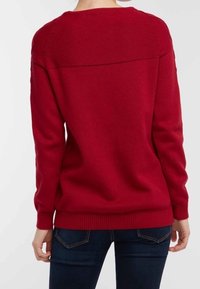 Roter Strickpullover mit rundem Ausschnitt, strukturiertem Stoff, gerippten Säumen und einfachem Design. Über dunklen Jeans getragen.