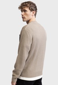 Maglione lavorato a maglia beige con un motivo a trama, polsini e vita a coste, e una vestibilità rilassata. Indossato sopra una camicia bianca.