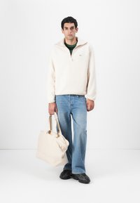 Light beige fleece pullover with a quarter zip, paired with blue flared jeans. The model holds a cream tote bag, wearing black shoes.