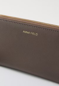 Brun læderpung med guldfarvet lynlås og prægget "ANNA FIELD" logo. Glat tekstur med en slank, rektangulær form.