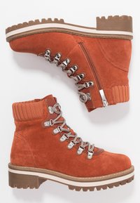 Botines de ante naranja con cuello acanalado texturizado, ojales de metal y cremallera frontal. Suela gruesa marrón con raya blanca y dibujo profundo.