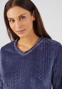 Top in morbido velluto blu navy con un motivo a righe testurizzate, dotato di scollatura a V e di un sottile accentuazione dorata attorno al collo. Materiale morbido e accogliente.