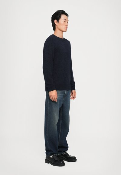Suéter texturizado azul marinho com gola redonda, combinado com jeans desbotados de corte largo e sapatos pretos robustos, visto de lado.