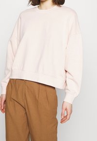 Ljust rosa sweatshirt med en boxig passform, rund halsringning och neddragna axlar, ihop med bruna vida byxor. Slät tygtextur.