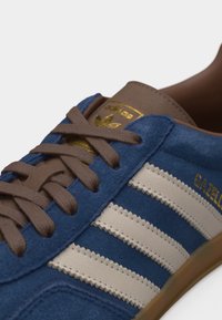 Μπλε σουέτ αθλητικό παπούτσι Adidas Gazelle με λευκές ρίγες, καφέ κορδόνια και χρυσή λεπτομέρεια λογότυπου στη γλώσσα και στο πλάι.