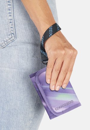 Hand holding a purple Coocazoo wallet with a black wrist strap, next to light blue denim jeans.