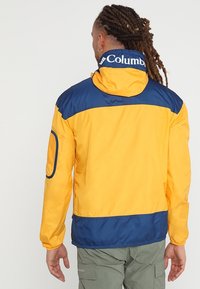 Chaqueta impermeable amarilla y azul marino con capucha, que cuenta con un bolsillo de malla en el brazo izquierdo y un logotipo a lo largo del cuello. Textura suave.