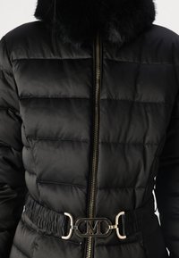 Chaqueta negra acolchada con cremallera dorada, cinturón elástico negro con hebilla dorada con la letra "M" y cuello de piel negro.