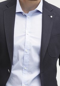 Blazer strutturato blu navy con un unico bottone, abbinato a una camicia azzurra con bottoni. Il blazer presenta una tasca sul petto e rever lisci.