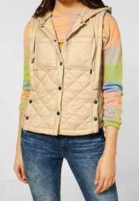 Beige prošívaná vesta s kapucí, obsahující přední kapsy na klopu, patentky a nastavitelné šňůrky. Nositá přes svetr s vícero barvami a pruhy.