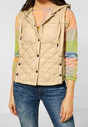 Beige quiltad väst med huva, som har framfickor med klaff, tryckknappar och justerbara dragsnören. Bärs över en randig tröja i flera färger.