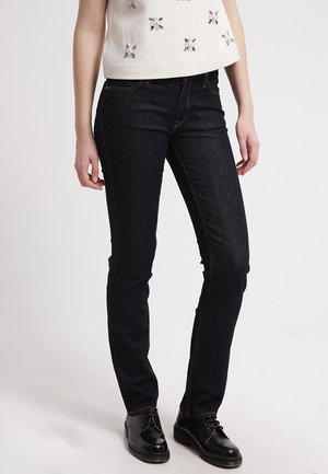 Mujer vistiendo jeans negros ajustados, zapatos negros con cordones y una camiseta sin mangas blanca con adornos decorativos.