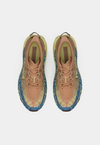 Os sapatos Hoka Speedgoat 6 apresentam um cabedal em malha bege com detalhes em azul, uma entressola texturizada e uma sola com padrão para tração.