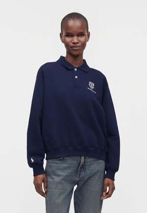 Person iført en mørkeblå langærmet polo-sweatshirt med Wimbledon-logo og lyseblå jeans, stående mod en ensfarvet baggrund.