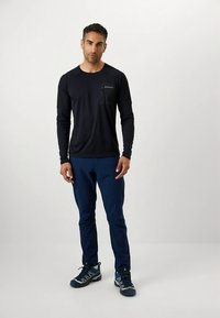 Camiseta preta de manga comprida com bolso no peito, combinada com calças azul-marinho e sapatos escuros com detalhes em azul, sobre um fundo claro.