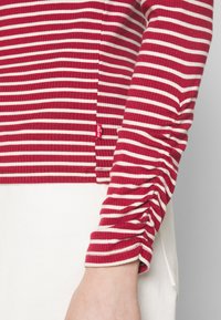 Chemise à manches longues à rayures horizontales rouges et blanches avec détail froncé sur les manches et petite étiquette de la marque Levi's, portée avec un pantalon blanc.