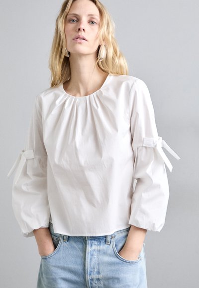 Fehér puffos ujjú blúz kerek nyakkivágással és ráncolt részletekkel, amelyen megkötők vannak az ujjak mandzsettáján, sima, ropogós pamutszövetből készült.