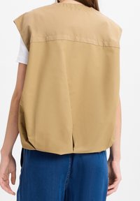 Beige ärmellose Weste mit glatter Textur, versehen mit einer Mittelnaht hinten und einem leicht gerafften Saum, kombiniert mit blauen Denim-Hosen.
