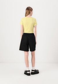 T-shirt jaune clair à manches courtes, short noir à taille élastique et sandales noires à plateforme. Cheveux lisses, avec un tatouage visible sur le bras.