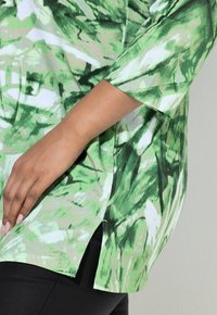 Zielony wzorzysty top z rękawami do połowy przedramienia. Wykonany z miękkiego materiału, zdobiony abstrakcyjnymi wzorami liści w różnych odcieniach zieleni i bieli. Z rozcięciami po bokach.