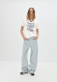 T-shirt blanc avec texte graphique et illustrations en bleu, associé à un jean large à rayures verticales bleu clair. Baskets de couleurs variées.