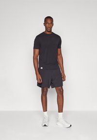 Camiseta negra de manga corta y pantalones cortos a juego. Hecho de una tela suave con una textura lisa. Lleva puesta con zapatillas blancas y calcetines tobilleros.