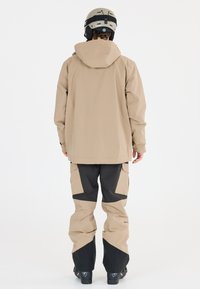 Beige vattentät jacka med kapuschong, matchad med svarta skidbyxor med stora fickor och snedställda färgblock. Svarta skidstövlar synliga.