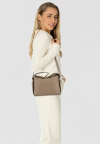 Borsa a spalla in pelle sintetica marrone con parte superiore arrotondata, tracolla regolabile e dettagli in argento, esposta su uno sfondo grigio chiaro.
