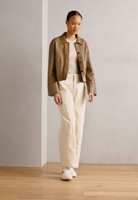 Giacca in pelle marrone sopra un top beige chiaro, abbinata a pantaloni a gamba larga color crema e sneakers bianche. Texture liscia e design casual.