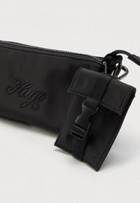 Μαύρη θήκη με φερμουάρ, με συνημμένη μικρότερη τσέπη με καπάκι, ασφαλισμένη με μαύρο πλαστικό κούμπωμα και λουρί, σε απλό φόντο.