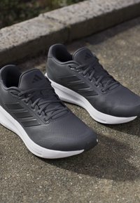 Zapatillas deportivas grises con un empeine texturizado, suelas blancas, cordones y tres rayas negras a cada lado. Diseñadas para la comodidad y el rendimiento.
