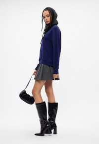 Kék kapucnis felső, szürke csíkos rakott szoknya, fényes fekete térd hosszúságú csizma és fekete kézitáska. Vízszintes és függőleges vonalak láthatók.