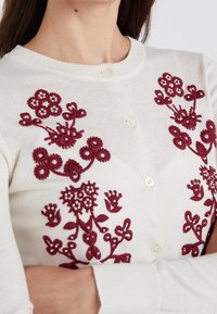 Valkoinen neulejakku, jossa on burgundinväristä kukkakirjontaa helmikoristeilla, pyöreä pääntie ja napitus. Silkkinen neulos, istuva leikkaus.