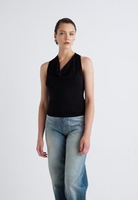 Schwarzes ärmelloses Oberteil mit einem Cowl-Ausschnitt, kombiniert mit hellblauen, leicht abgetragenen, geraden Jeans. Minimalistisches Design mit glatter Textur.