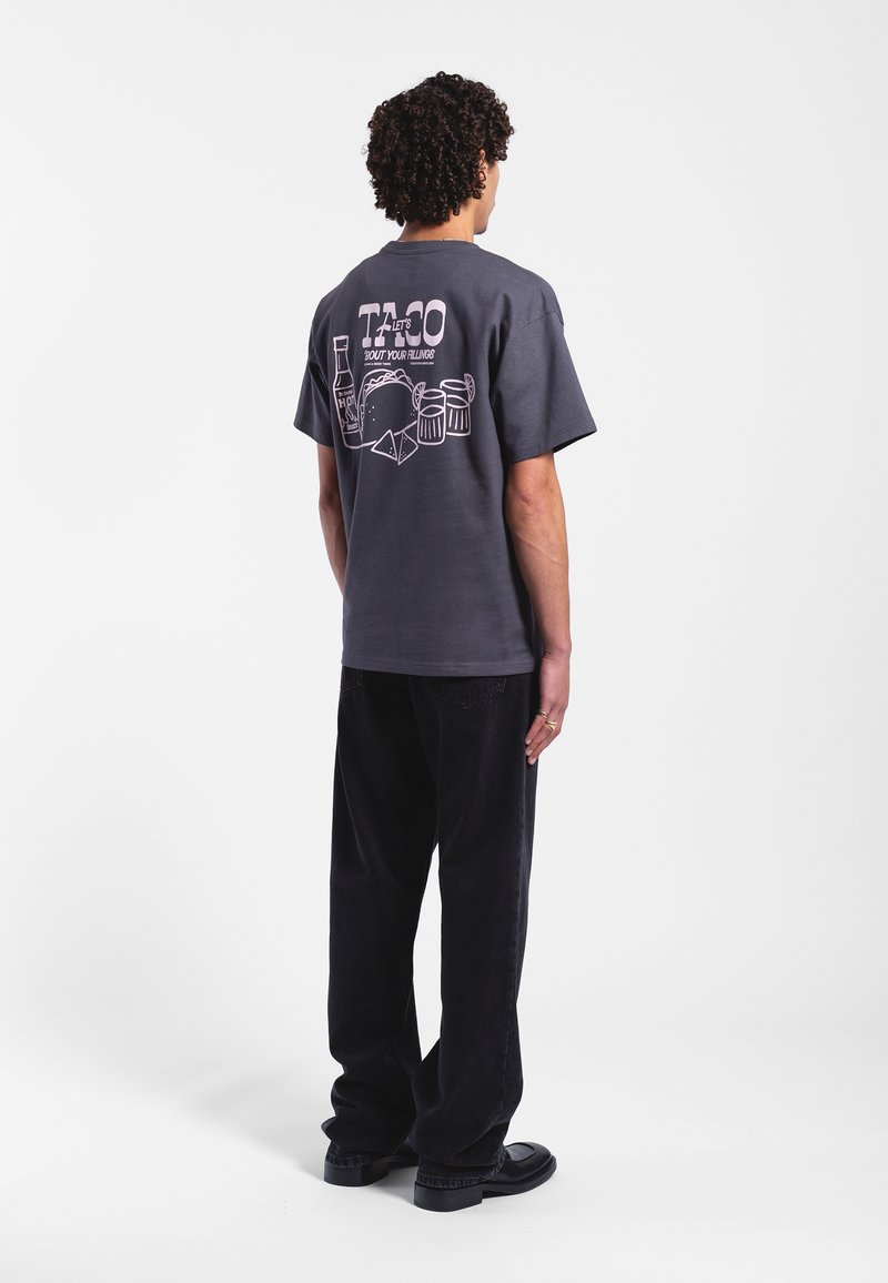 Grå bomullströja med korta ärmar har en grafisk tryck på baksidan som föreställer en taco, drycker och text. Kombinationen med svarta byxor.