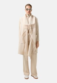 Béžový šál z vlněné směsi s třásněmi na okrajích, doplněný o béžový outfit ve stejném odstínu a kulaté sluneční brýle. Hladká textura a minimalistický design.