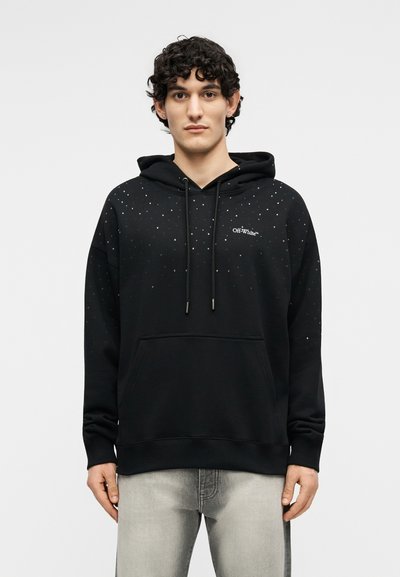 Jauns vīrietis, kas vērsts uz priekšu, valkā melnu Off-White kapuci ar mazām baltām punktiem un gaiši pelēkas džinsas, fonā ir vienkrāsains fons.