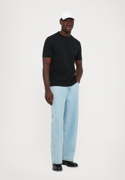 Junger Mann trägt ein schwarzes kurzärmliges Hemd, helblaue weit geschnittene Jeans, schwarze Schuhe und eine weiße Baseballkappe und steht vor einem schlichten Hintergrund.