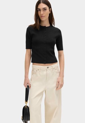 Mujer con una blusa negra de manga corta y botones, y pantalones anchos de color crema, sosteniendo un bolso negro en la mano izquierda.