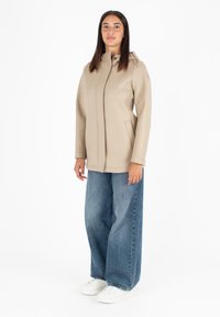 Cappotto in misto lana beige con colletto alto, tasche laterali e taglio dritto, abbinato a jeans in denim blu larghi e sneakers bianche.