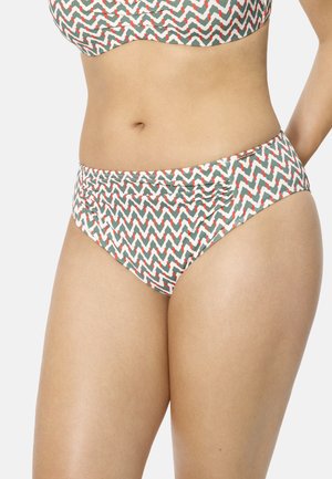 Mujer con braguitas de bikini de talle alto con patrón de zigzag verde y rojo y top de bikini a juego, de pie con un brazo doblado hacia atrás.