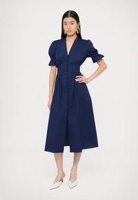 Μίντι φόρεμα σε ναυτικό μπλε με φουσκωτά μανίκια, ντεκολτέ σε σχήμα V και κούμπωμα με κουμπιά στο μπροστινό μέρος. Ομαλή υφή και εφαρμοστό μπούστο με φούστα σε γραμμή καμπάνα.