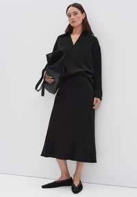 Blouse noire avec un décolleté en V, associée à une jupe noire longueur cheville. Les accessoires comprennent un sac en maille et des ballerines noires avec des accents cloutés.