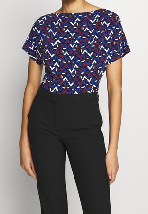 Femme portant un haut à manches courtes avec un motif géométrique bleu, blanc et rouge rentré dans un pantalon noir taille haute.