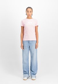 Roza pamučna majica s kratkim rukavima i bijelim logotipom. Uparena s svijetloplavim širokim trapericama i bijelim tenisicama. Bijela pozadina.