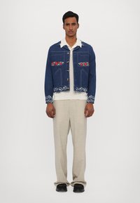 Denim jakna v temno modri barvi z rdečim cvetličnim vezenjem na žepih in manšetah, zlatimi gumbi, skrajšano zasnovo, nosena čez belo srajco in bež hlače.