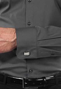 Camicetta grigia con una leggera lucentezza, dotata di polsini con bottoni e gemelli. Il tessuto è liscio con una leggera texture, abbinato a una cintura nera.