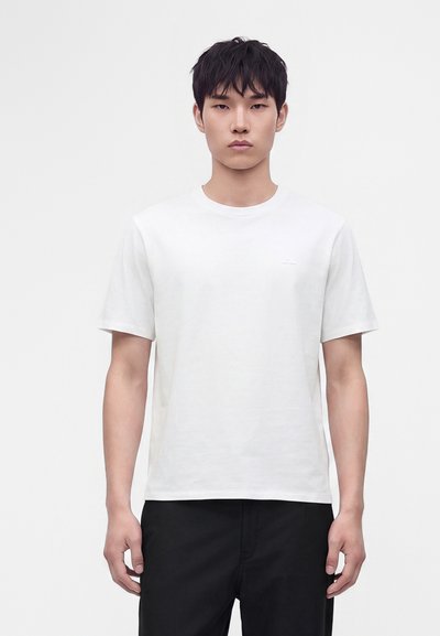 Jeune homme aux cheveux noirs courts portant un t-shirt blanc uni à manches courtes et un pantalon noir, debout devant un fond blanc.
