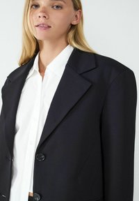 Czarna marynarka o strukturalnym fasonie, z szerokimi klapami i dwoma guzikami. Noszona na białej koszuli z kołnierzykiem, co podkreśla klasyczny, dopasowany wygląd.